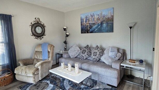 Un salón con un sillón color beige, un sofá gris, una mesa de centro blanca y un cuadro de un paisaje urbano.