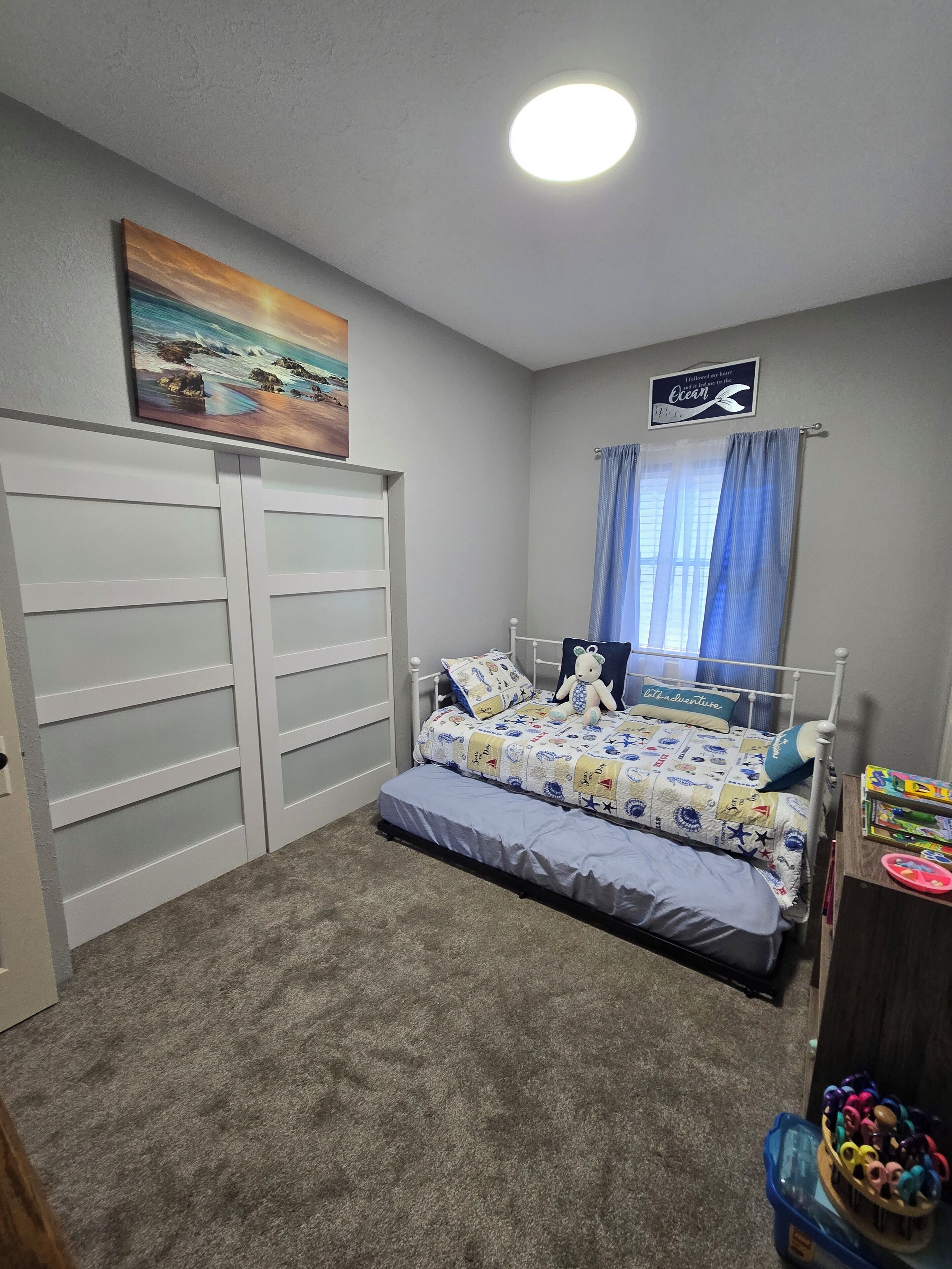 A bedroom with a bed, a dresser, a painting, a window with curtains, and a sign on the wall.