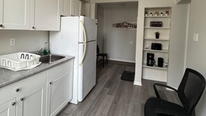 A compact kitchen with white cabinets, a white refrigerator, and a black office chair. - Serenity Suite - 2Br (Cornwall)