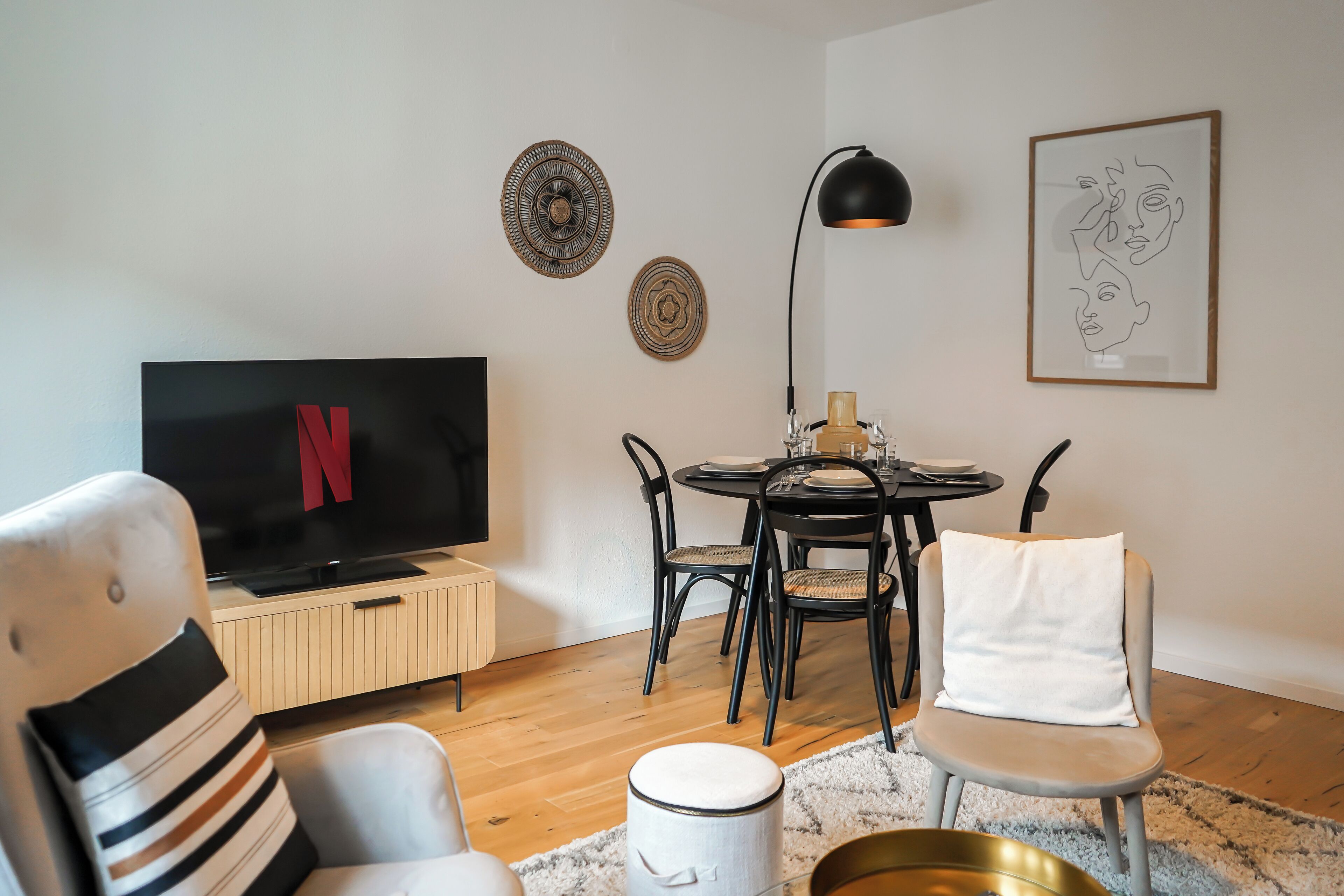 A modern living room with a flat-screen TV, a dining table with chairs, a lamp, and decorative wall art.