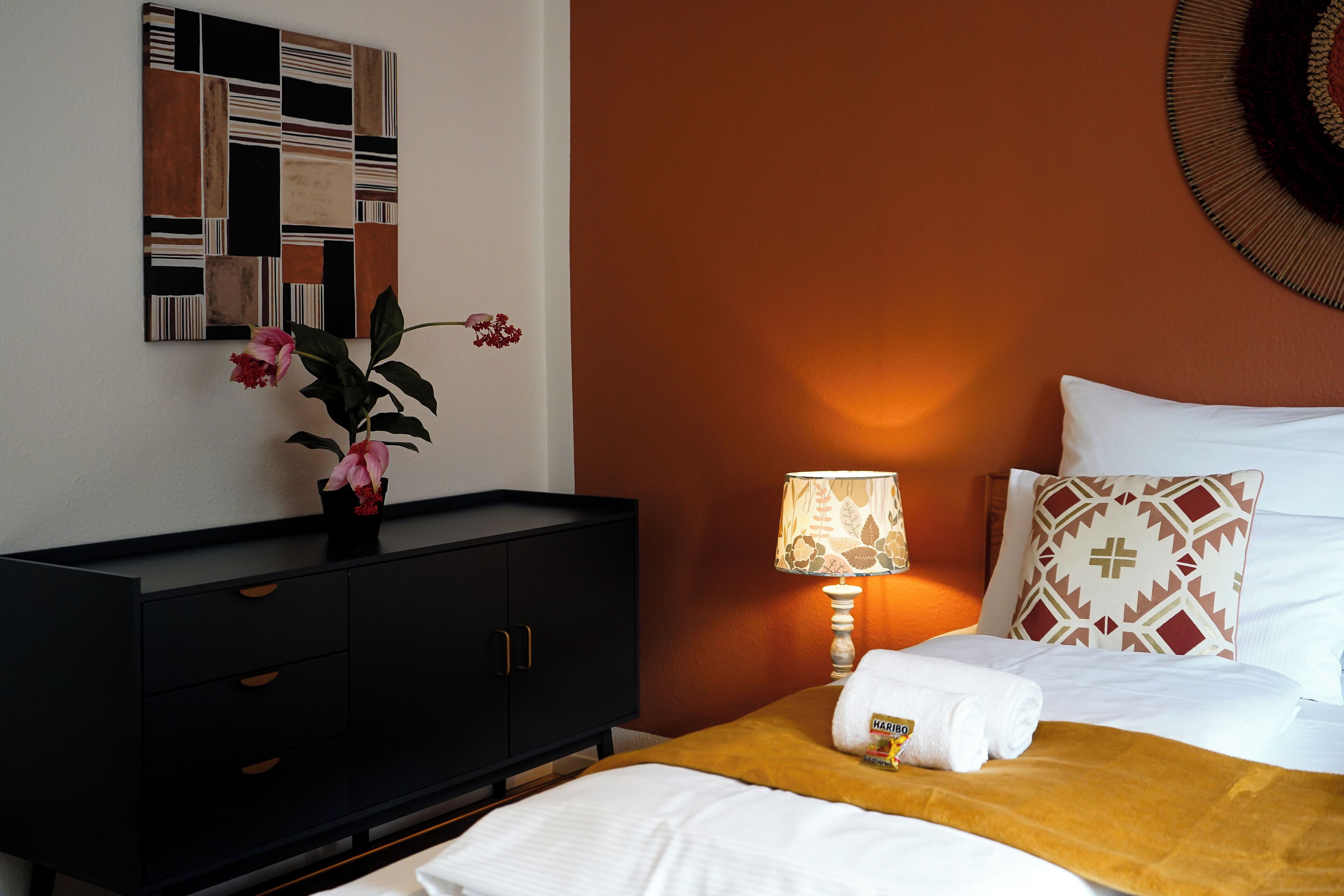 Quarto de hotel com uma cama, mesa de cabeceira com abajur, um objeto decorativo na parede e um vaso com planta.