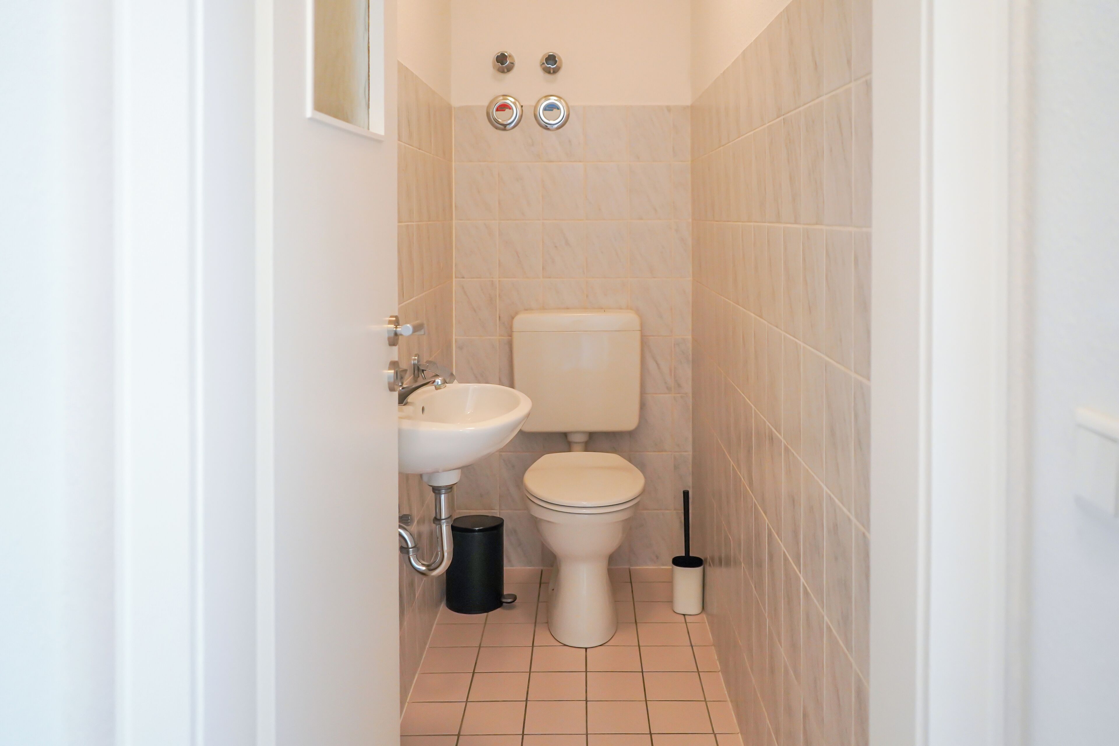 A small, clean bathroom with a toilet, sink, and two wall-mounted flush valves.