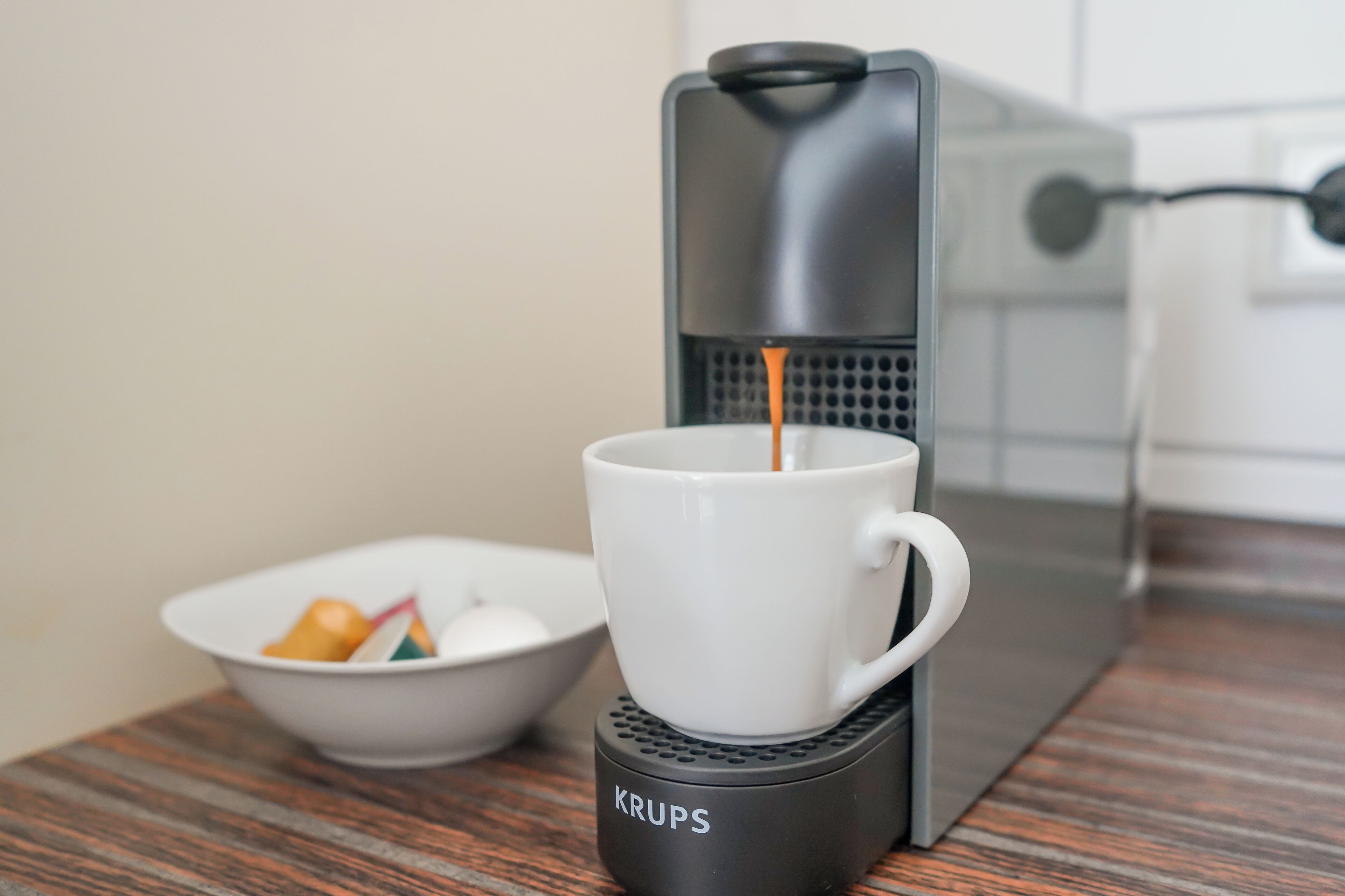Una cafetera sirviendo café en una taza blanca, con una cafetera Krups y un tazón de cubitos de azúcar y bolsitas de té sobre una mesa de madera.