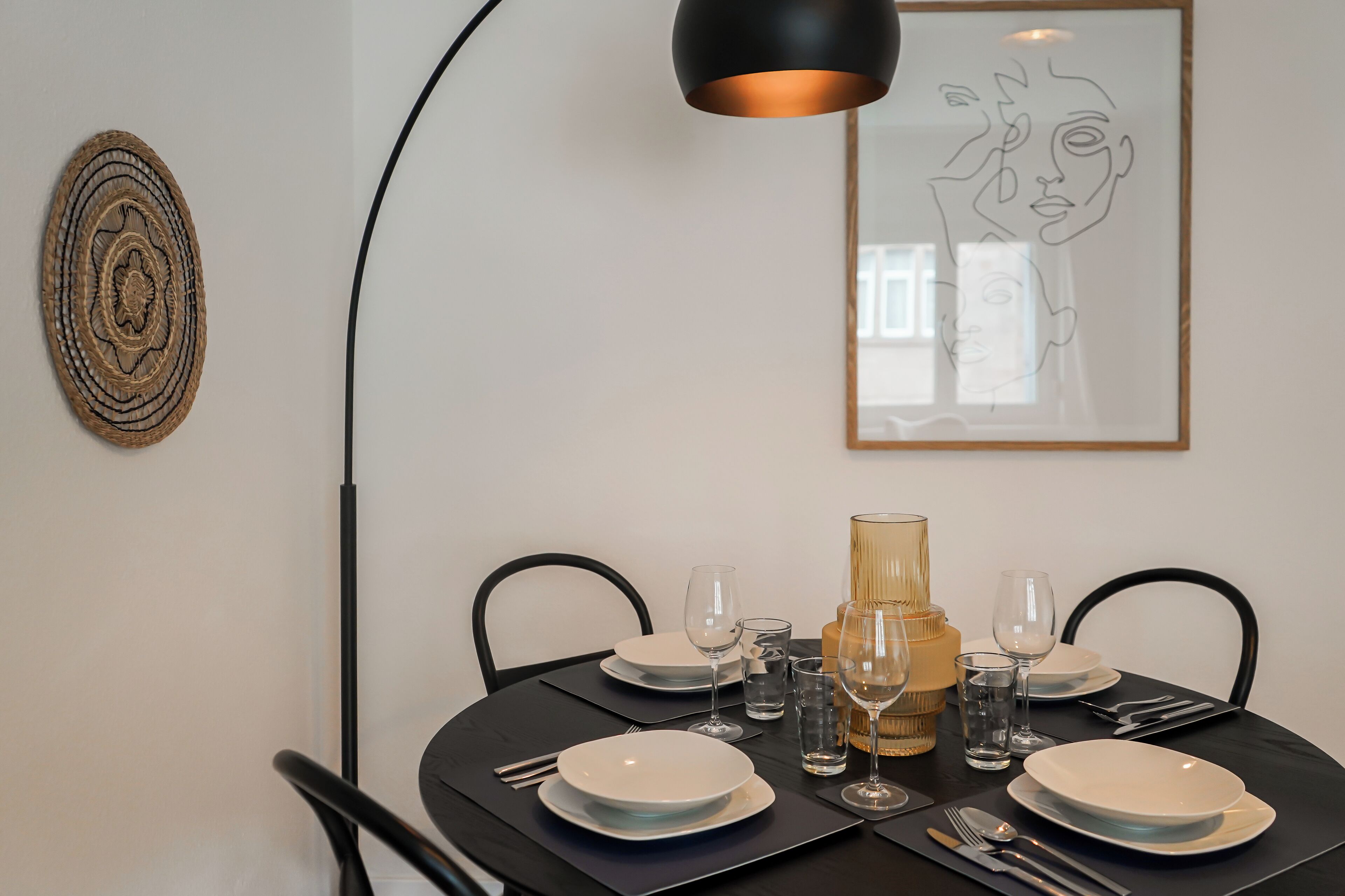 Mesa de jantar posta com pratos, copos e talheres, uma luminária e uma obra de arte na parede.