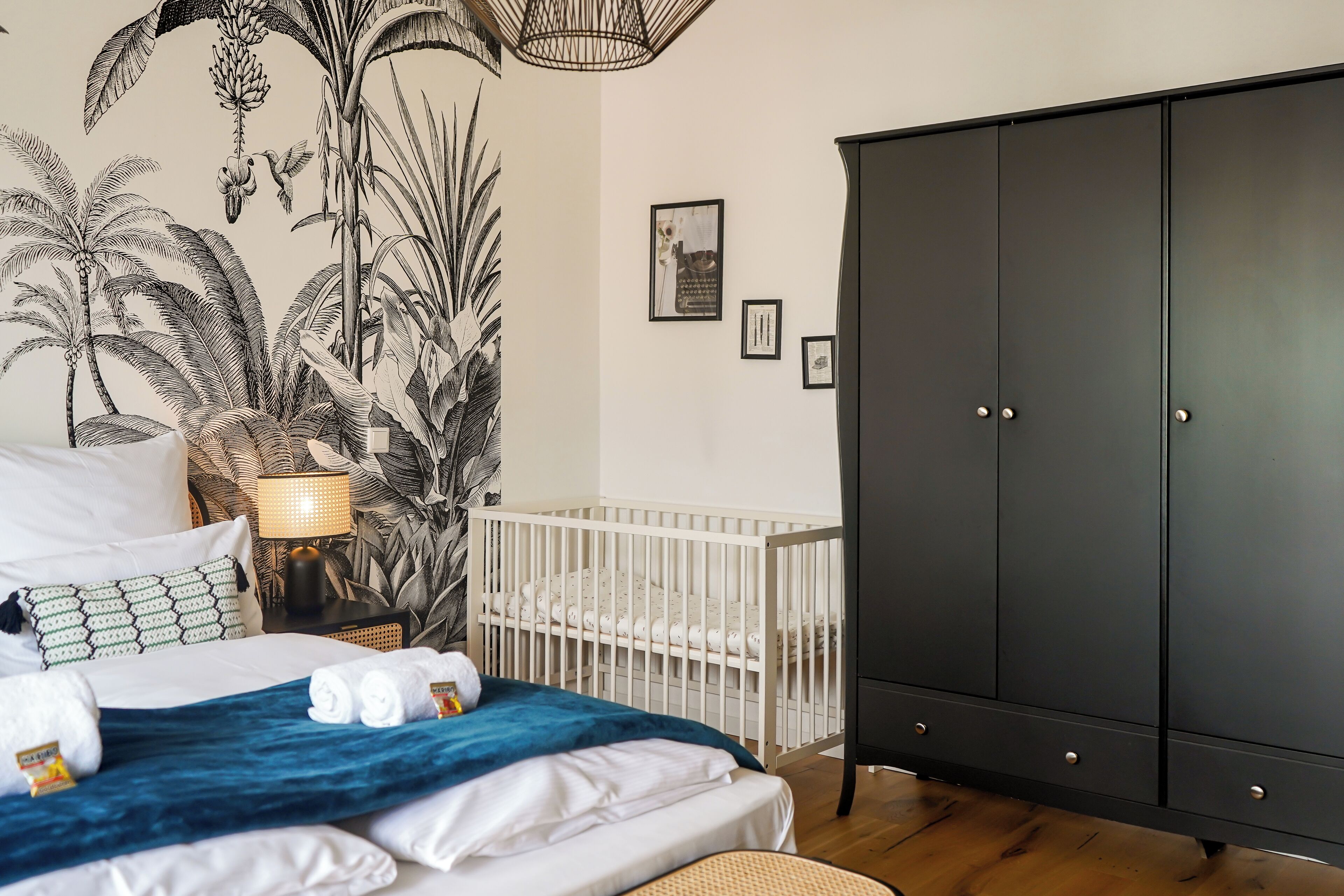 Quarto com berço, guarda-roupa escuro, cama com roupa de cama branca e manta azul, e um mural de plantas tropicais na parede.