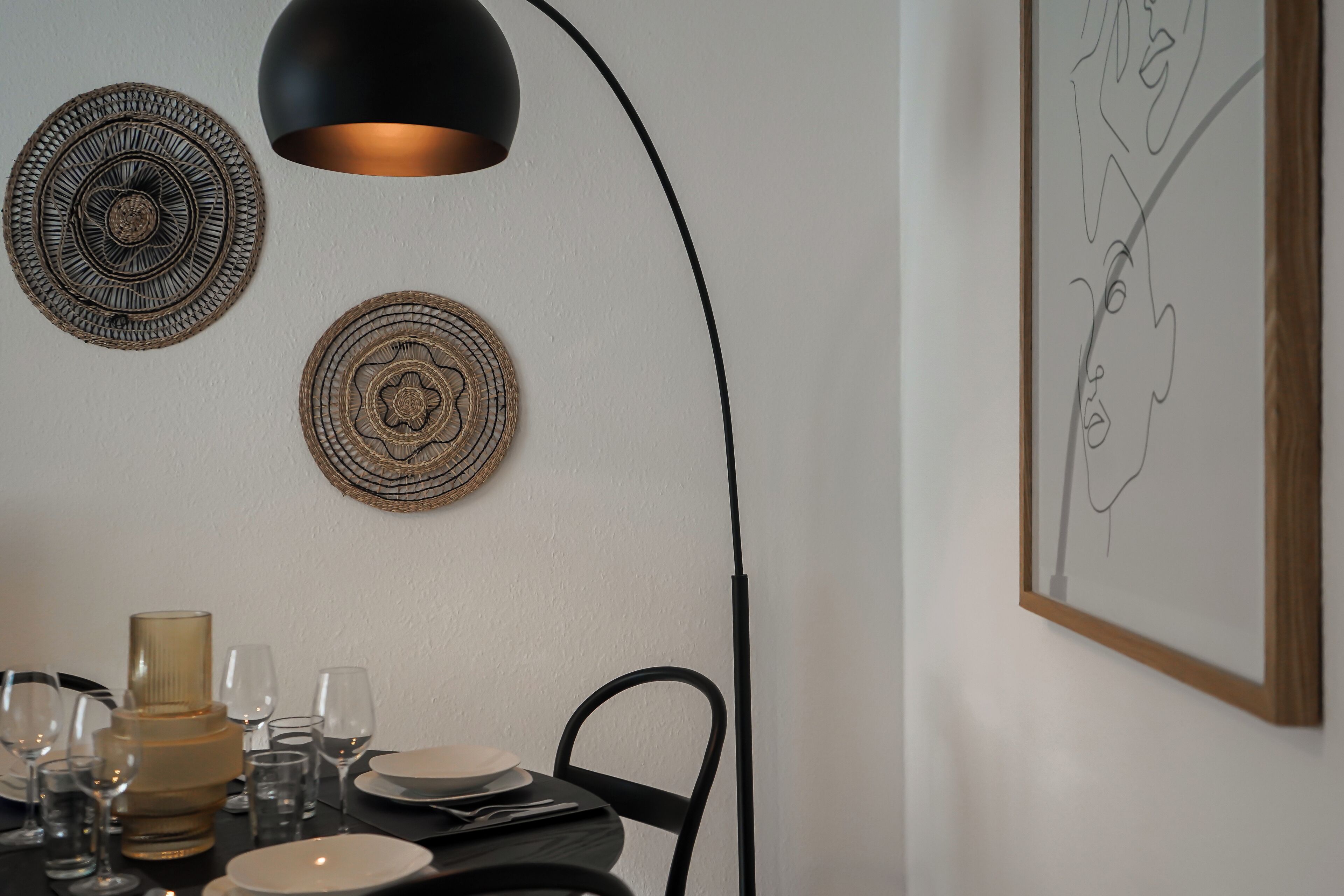 Een eetgedeelte met een tafel voor twee personen, met een zwarte stoel, een zwarte hanglamp en twee wandplaten van geweven materiaal.