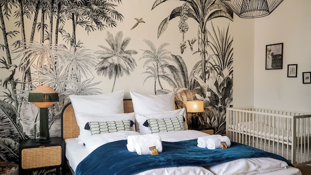 A bedroom with a large bed, a crib, a nightstand, a lamp, and a wall mural of tropical plants.