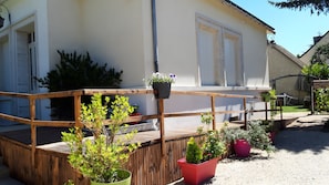 Une terrasse en bois agrémentée de plantes en pot, un bâtiment blanc avec des volets et un ciel dégagé.