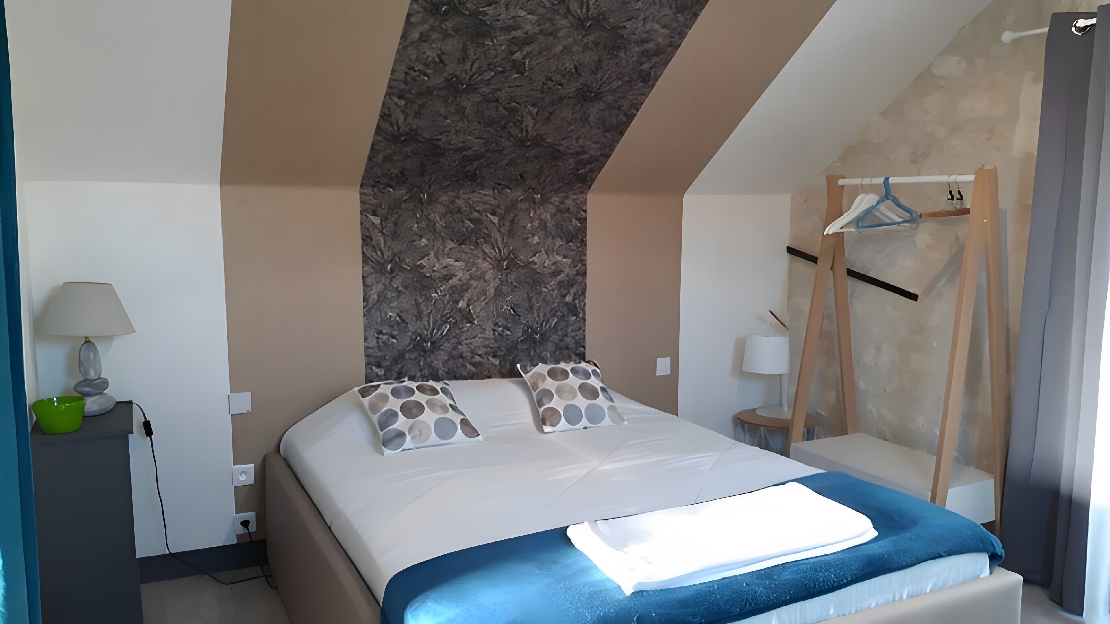 A hotel room with a bed, pillows, a lamp, a bowl, a hanging rack, and a wall with a textured pattern.