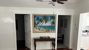 Ein geräumiges Zimmer mit Holzboden, Deckenventilator, einem Strandbild und einer Holzbank.