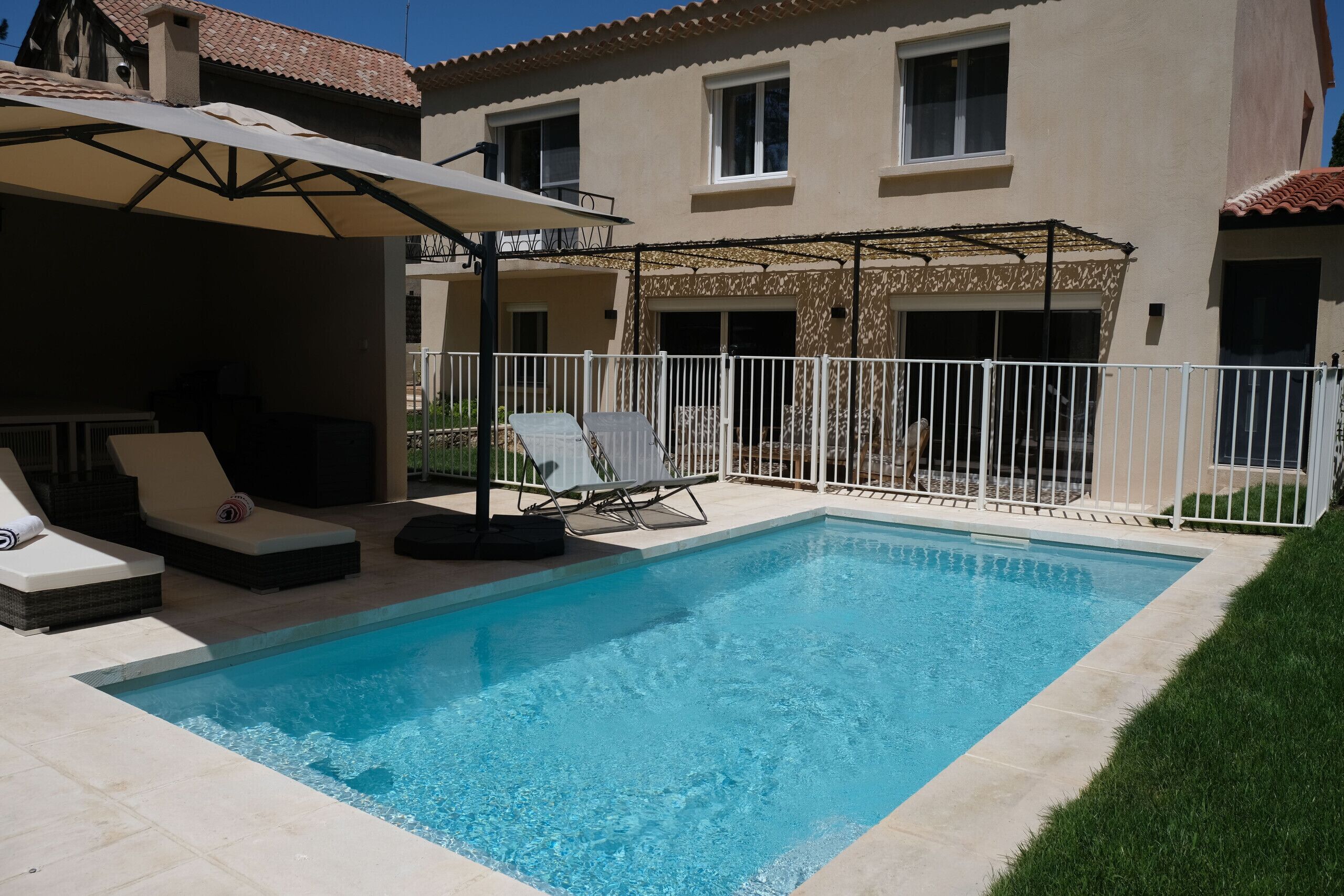 A rectangular swimming pool with lounge chairs and umbrellas, surrounded by a white fence and a building with multiple windows.