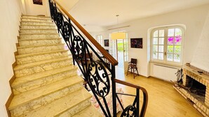 Un escalier en marbre, agrémenté de rampes en fer noir, mène à une pièce dotée d’une cheminée et d’une grande fenêtre offrant une vue sur la verdure.