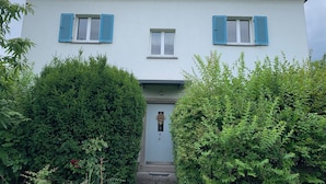 Een gebouw met twee verdiepingen, een witte gevel en blauwe luiken, omringd door weelderige begroeiing.
