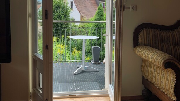 Een open deur die leidt naar een balkon met een tafel en stoel, een houten vloer binnen en een bank.