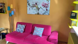 Een felroze bank met patroon kussens, een glazen salontafel en een lijst met een foto van een kolibrie op een perzikkleurige muur.