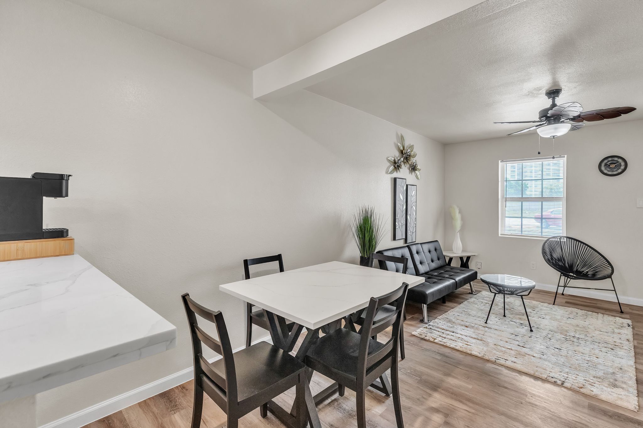 Un coin repas avec une table et des chaises blanches, un ventilateur de plafond, une fenêtre avec des stores et un canapé en cuir noir.
