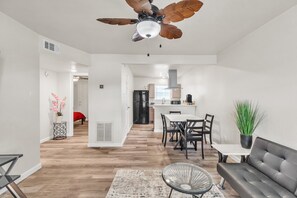 A modern living room with a ceiling fan, a dining area with a table and chairs, and a kitchen in the background.