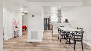 Una cocina moderna con una isla central, un comedor con una mesa blanca y sillas oscuras, y un pasillo con una cama roja y una planta decorativa.