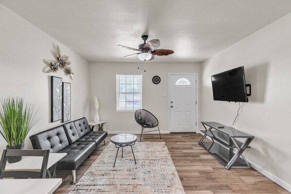 A living room with a black leather sofa, a round coffee table, a black chair, a white door, a window with blinds, a ceiling fan, and a flat-screen TV mounted on the wall.