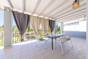A covered outdoor patio with a table and chairs, a view of greenery, and a railing.