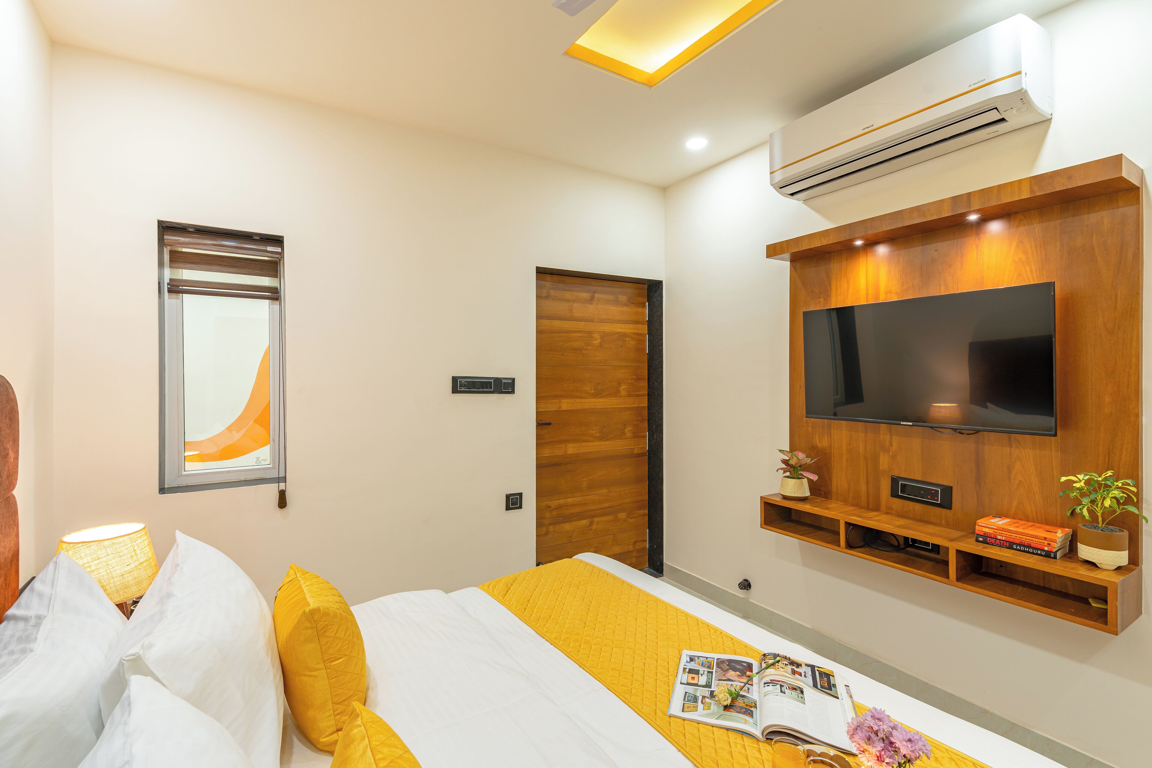 Quarto de hotel moderno com uma cama grande, uma TV de tela plana fixada em um painel de madeira, um ar condicionado e uma pequena estante com objetos decorativos.