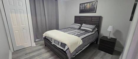 Een slaapkamer met een bed, nachtkastje, een deur en een schilderij aan de muur.