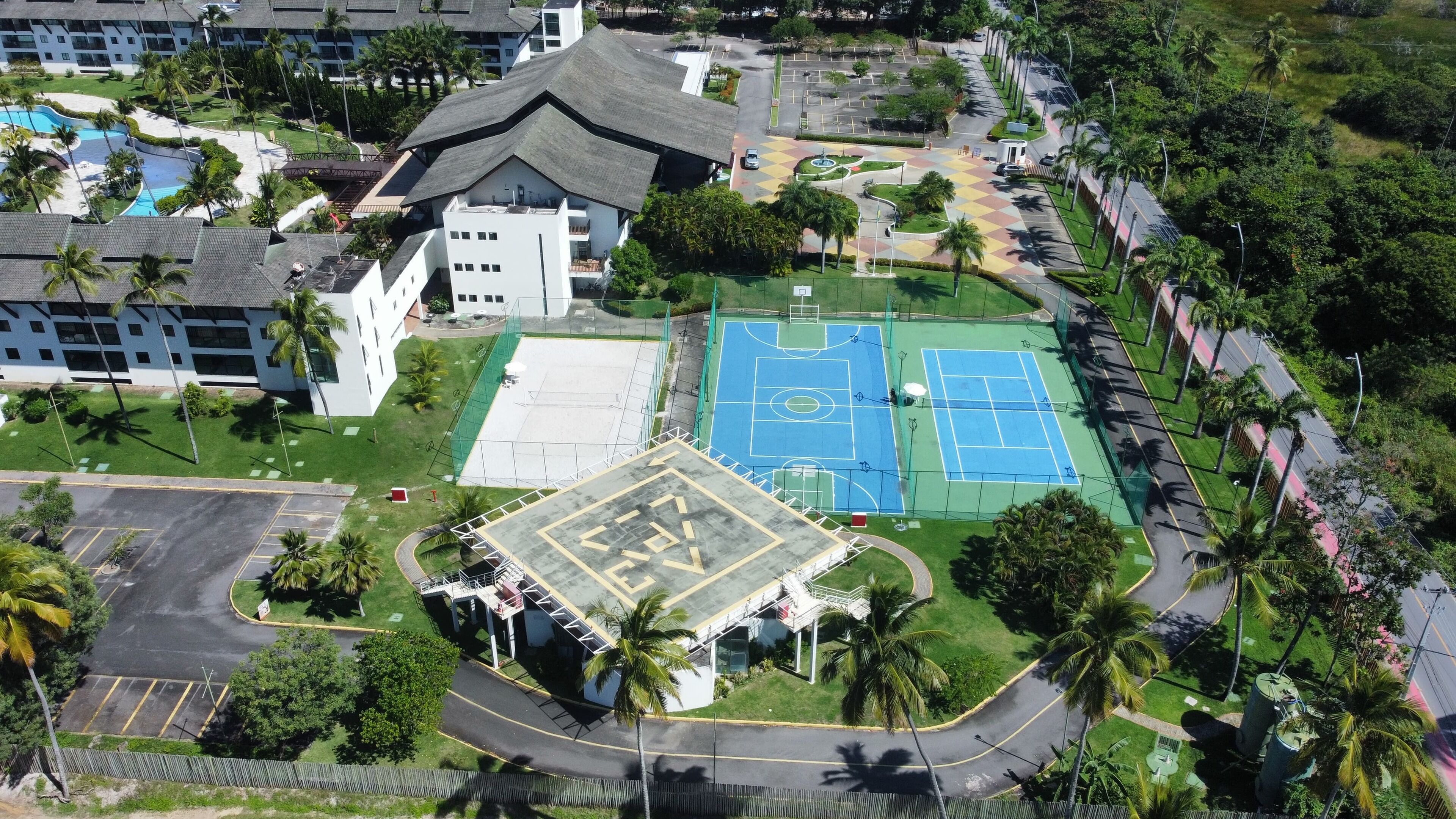 Vista aérea de um resort com piscina, quadras de tênis e um edifício com telhado inclinado.