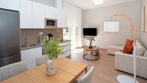 A modern kitchen with white cabinets, a stainless steel refrigerator, and a microwave. There is a dining area with a wooden table and chairs, and a living room with a sofa and a floor lamp.