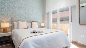 A neatly made bed with a white bedspread and two pillows, a bedside lamp on each side, and a window with blinds.