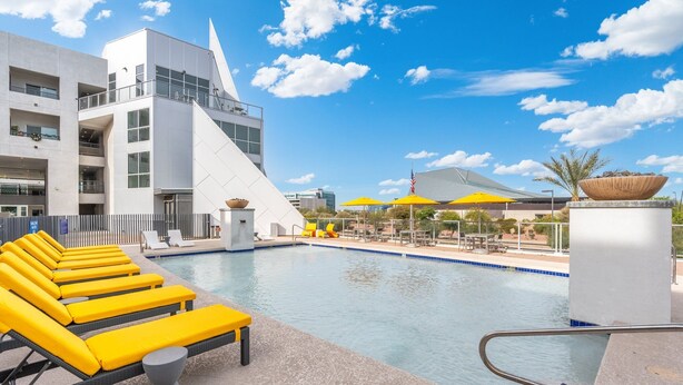 Ett modernt hotellområde med en pool, gula solstolar, en klarblå himmel och en distinkt byggnad med en triangulär design.
