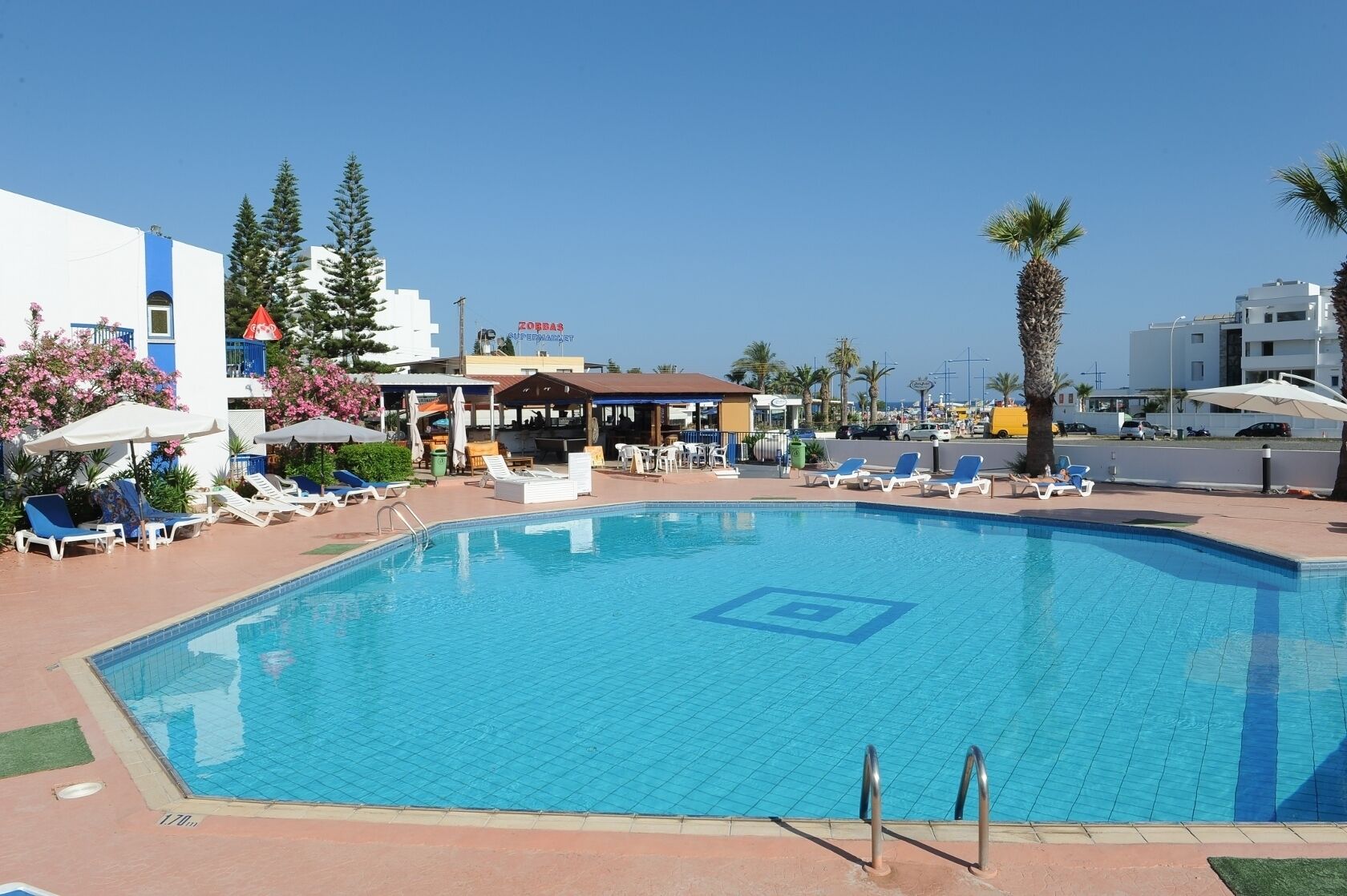 Una piscina con sillas de descanso y sombrillas, rodeada de edificios y palmeras.