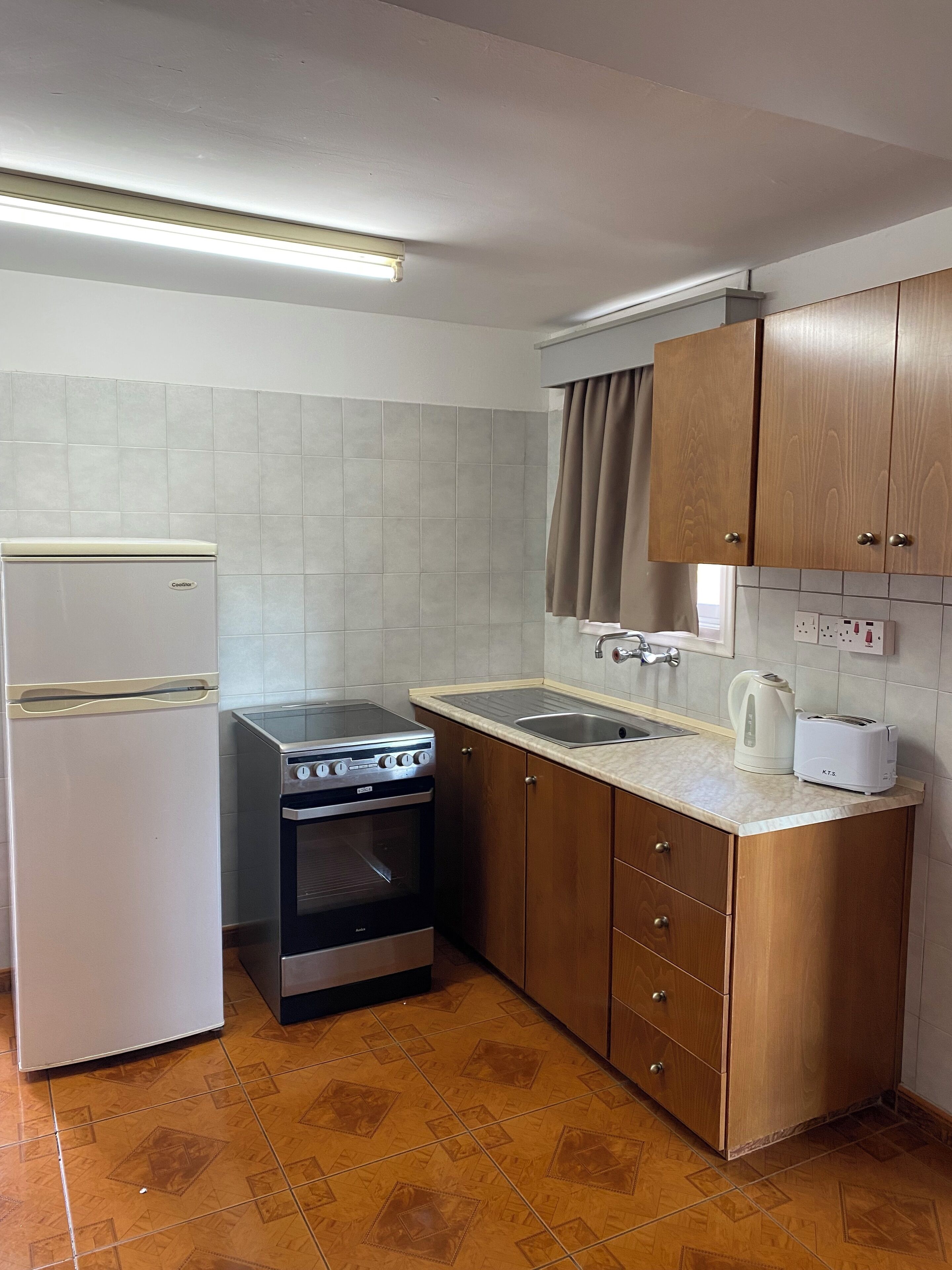 A compact kitchen with a refrigerator, stove, oven, and a sink with a kettle and toaster.
