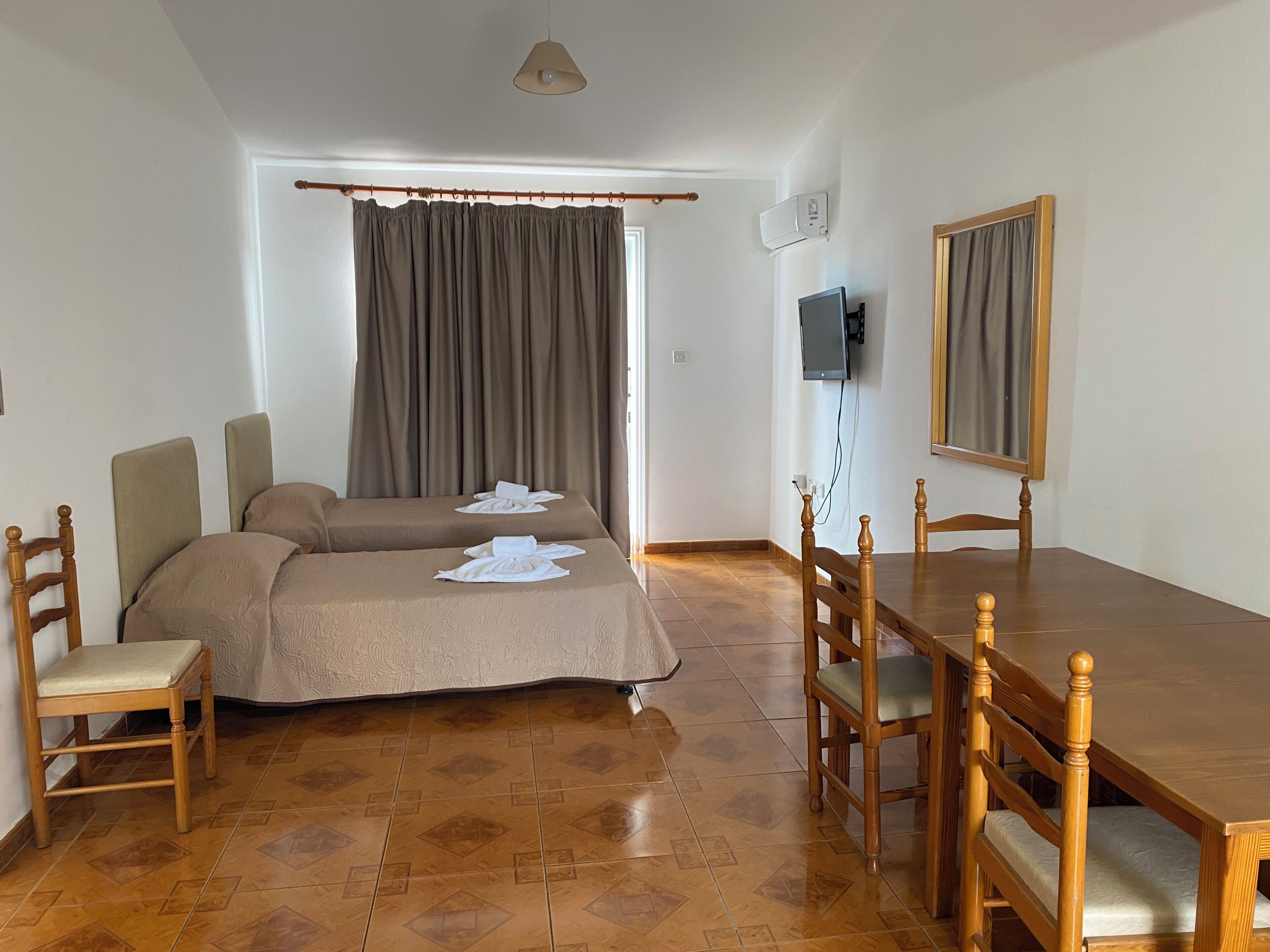 Een kamer met twee bedden, een houten eettafel, stoelen, een televisie en een raam met gordijnen.