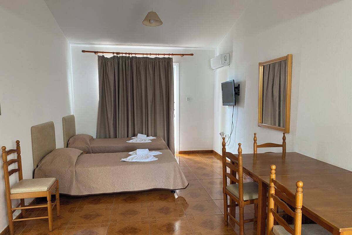 Ein Zimmer mit zwei Betten, einem Holztisch, Stühlen, einem Fernseher und einem Fenster mit Vorhängen.