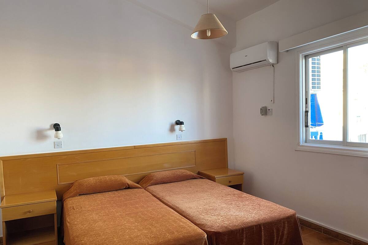 Ein Schlafzimmer mit hölzernem Kopfteil, zwei Nachttischen, einem Bett mit braunen Bettbezügen, einer Klimaanlage und einem Fenster mit Blick auf Gebäude.