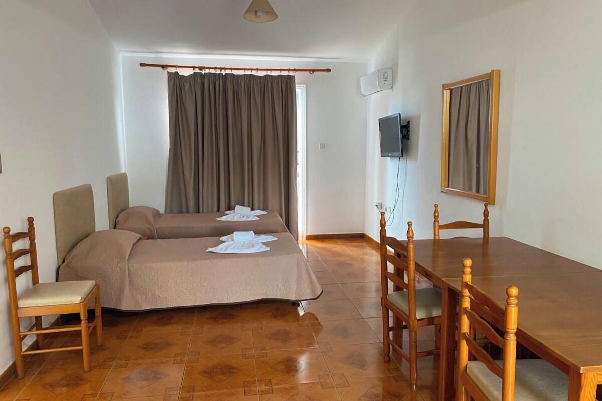 Ein Hotelzimmer mit zwei Betten, einem Holzesstisch, Stühlen, einem an der Wand montierten Fernseher und einem Fenster mit Vorhängen.