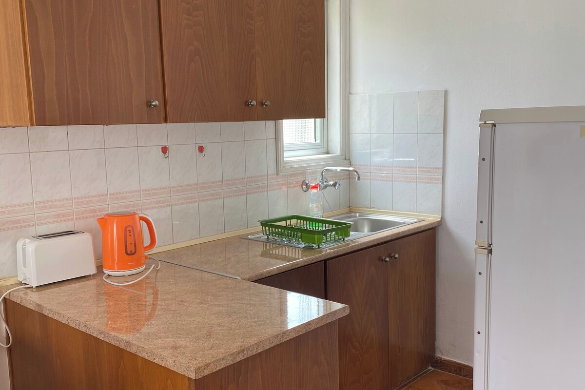 Een keuken met houten kastjes, een aanrecht, een broodrooster, een waterkoker, een spoelbak en een koelkast.