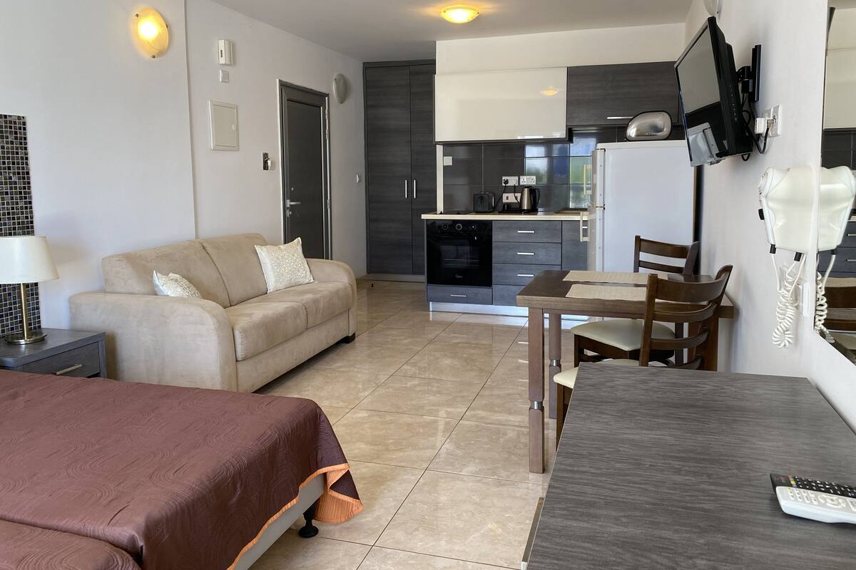 Ein modernes Hotelzimmer mit einer Sitzecke, einem Esstisch mit Stühlen, einer Küchenzeile inklusive Kühlschrank, Herd und Mikrowelle sowie einem an der Wand montierten Fernseher.