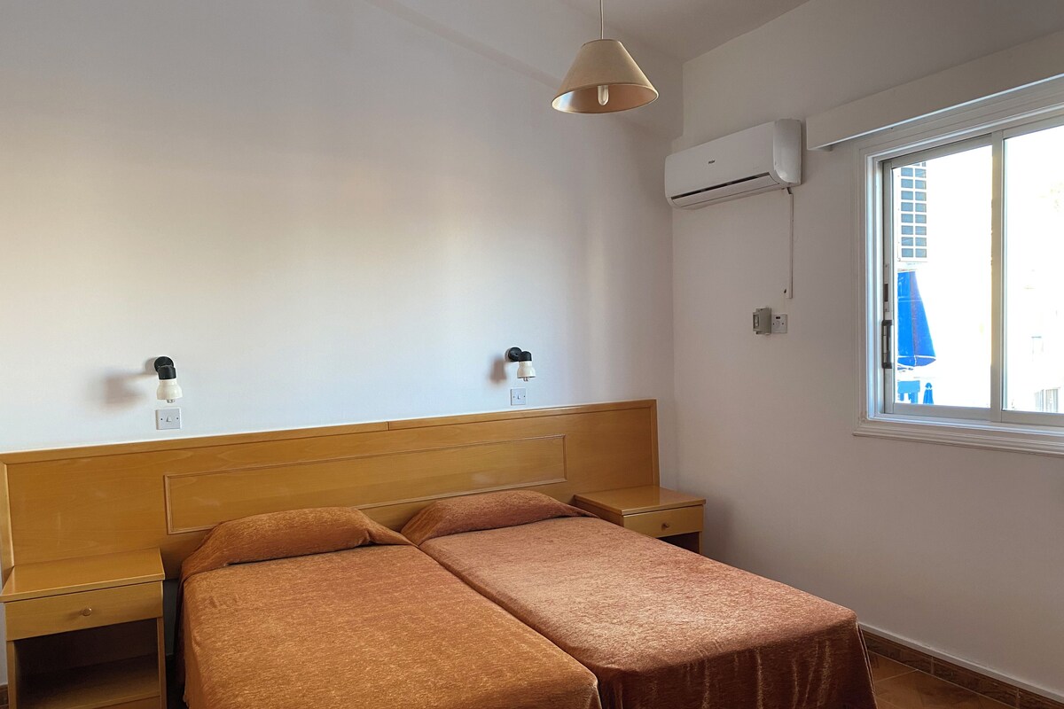 Ein Schlafzimmer mit Holzkopfende, zwei Nachttischen, einem Bett mit brauner Bettwäsche, einer Klimaanlage und einem Fenster mit Blick auf Gebäude.