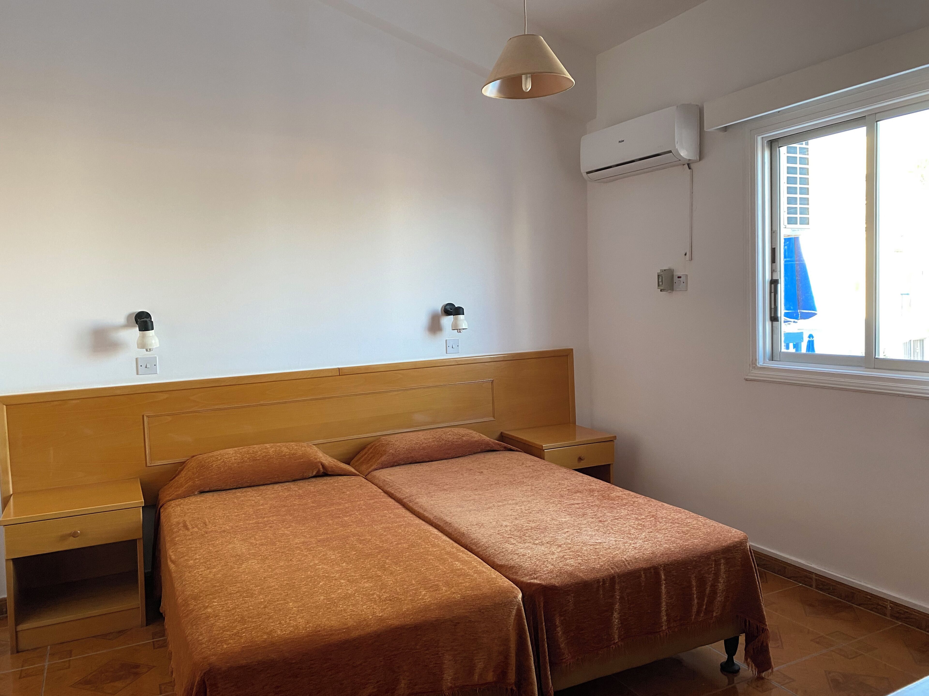 Een slaapkamer met een houten hoofdbord, twee nachtkastjes, een bed met bruin beddengoed, een airconditioner en een raam met uitzicht op gebouwen.