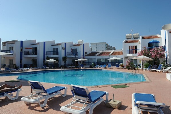 Une piscine entourée de bâtiments à plusieurs étages dotés de balcons, avec des chaises longues et des parasols à proximité.