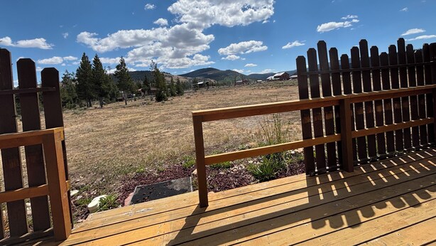 A wooden deck with a view of a grassy field, trees, and distant mountains under a partly cloudy sky.