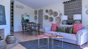 A living room with a sofa, two side tables, a dining table with chairs, and a kitchen area in the background.
