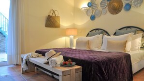 A bedroom with a bed, pillows, a bedside table with a bowl of fruit, a lamp, and a hanging basket.