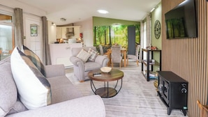 Ein Wohnzimmer mit Sofa, Couchtisch, Fernseher und Kamin. Im Hintergrund ist eine Küche mit grüner Wand und Essbereich zu sehen.