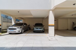 A covered parking area with two cars parked, a motorcycle, and a building with a white facade and yellow lines on the floor. - Apto bem Localizado a 500mts da Praia Rhi206 (Florianópolis)