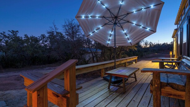 A wooden deck with a table and benches, an umbrella with string lights, and a building in the background. - Bison View Secluded Cabin for 6 w/ Hot Tub on Wildlife Sanctuary (Johnson City)
