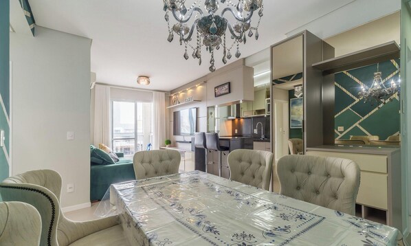 A modern living room with a dining table, chairs, and a chandelier. There is a kitchen area with a refrigerator and a dining table.
