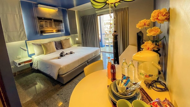 A hotel room with a bed, bedside table, lamp, and a dining area with a table, chairs, and a vase of flowers. - Room-hotel With Balcony in Phukettown (Phuket)