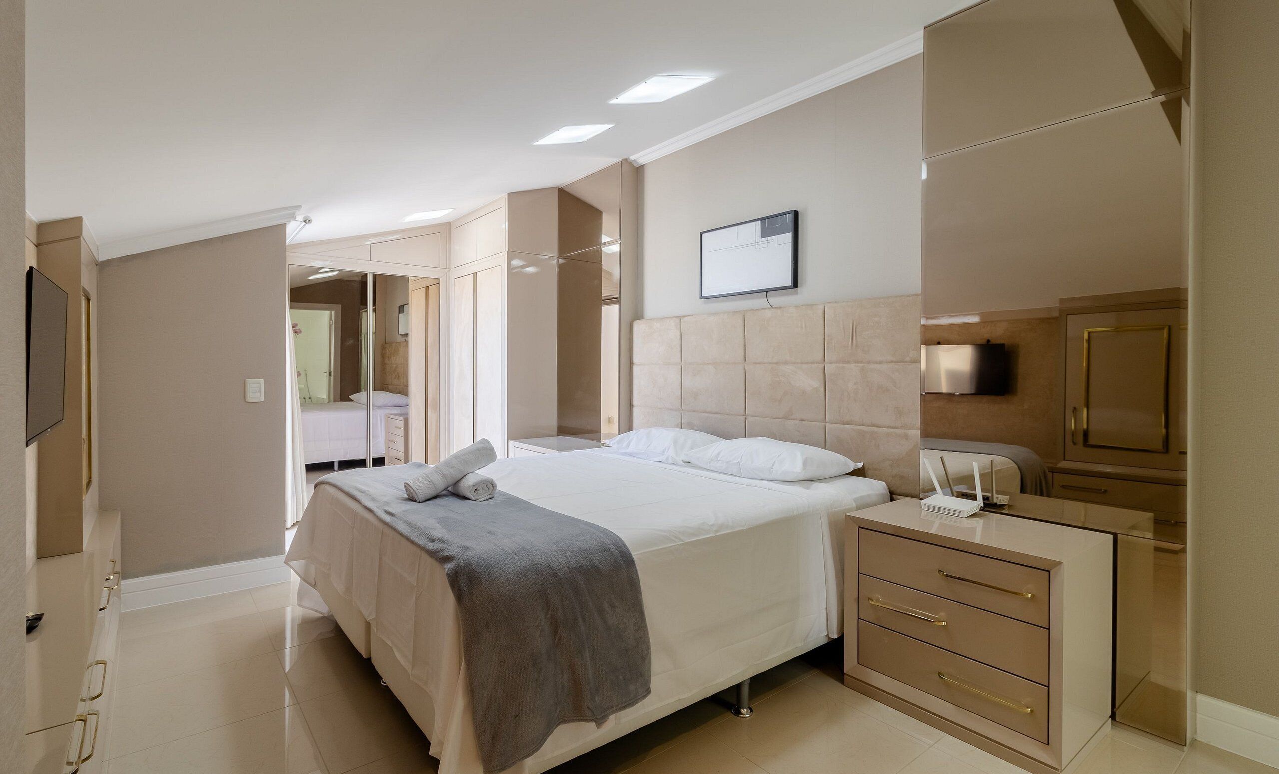 A modern hotel room with a large bed, a bedside table with a lamp, a wardrobe, and a flat-screen TV mounted on the wall.
